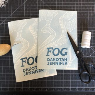 Two copies of Fog by Dakotah Jennifer overlapping on a black gridded cutting mat. The hand-printed linocut cover design consists of white curved lines in a wavelike vertical pattern on a light blue-gray background. Dark blue lettering spells out the title and author name in the lower center. Next to the books are some binding tools: a spool of linen thread, a pair of black scissors, and a wooden-handled awl.
