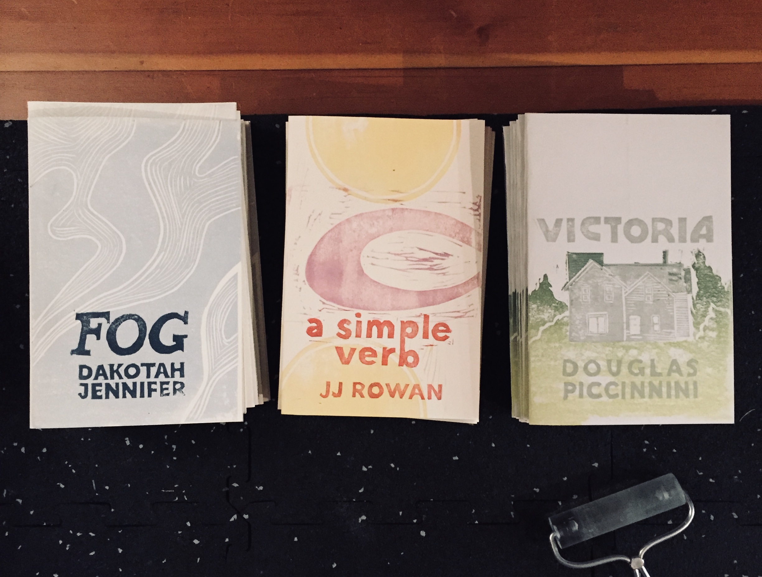 Three chapbooks with linocut covers. From left to right: FOG by Dakotah Jennifer, blue and cream waves with dark blue text; A SIMPLE VERB by JJ Rowan, yellow and lavender geometric shapes with red-orange text; and VICTORIA by Douglas Piccinnini, light green-to-dark green gradient ground with a gray house and gray text.
