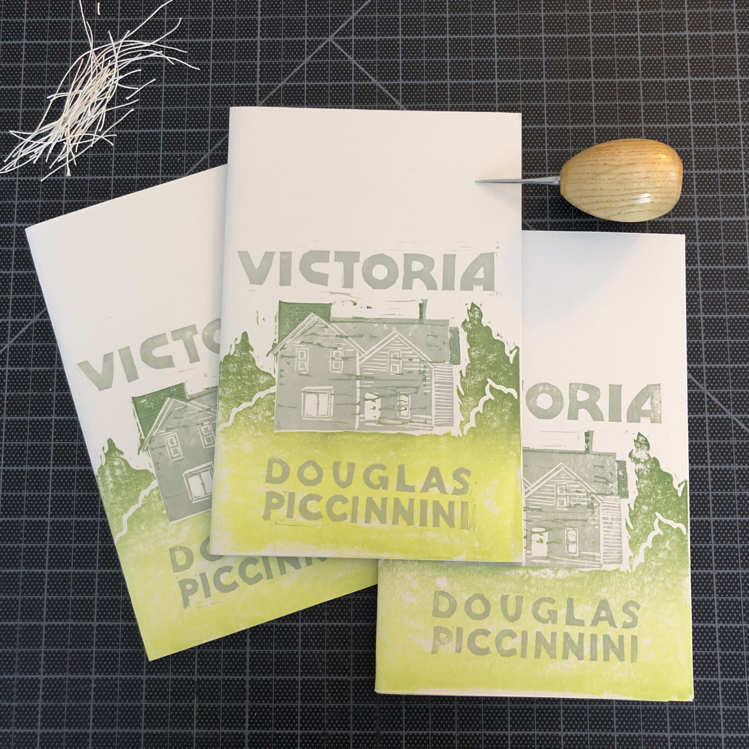 Three copies of Victoria by Douglas Piccinnini. The hand-printed linocut cover design consists of a gray house in a gradient green landscape, with gray title and author name lettering. The chapbooks are fanned out on a black gridded cutting mat, with a pile of string in the upper left, and a wooden-handled awl in the upper right.