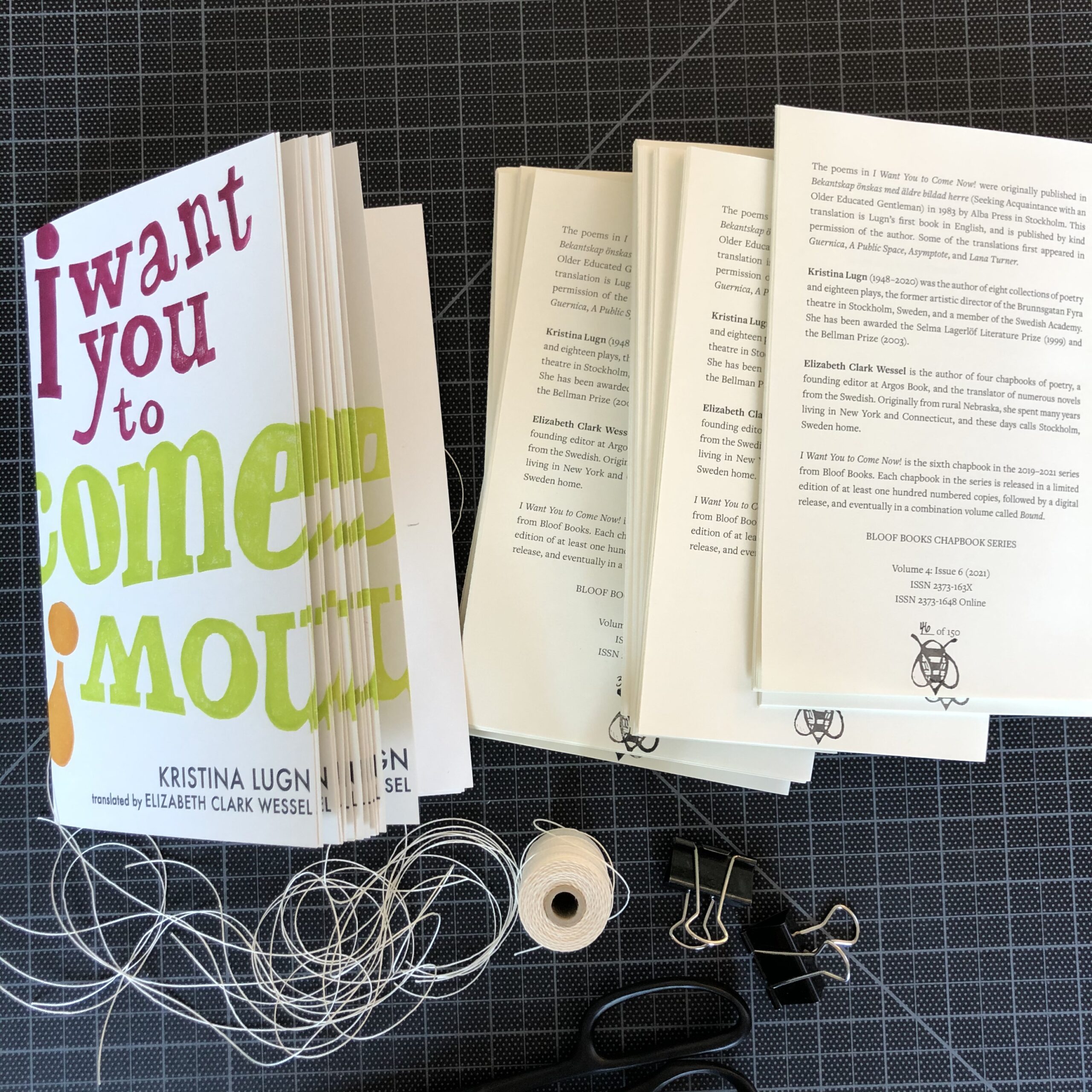 Copies of I Want You to Come Now! in progress. A stack of folded covers and a stack of folded interiors sit side by side on a black gridded cutting mat, with thread and binder clips