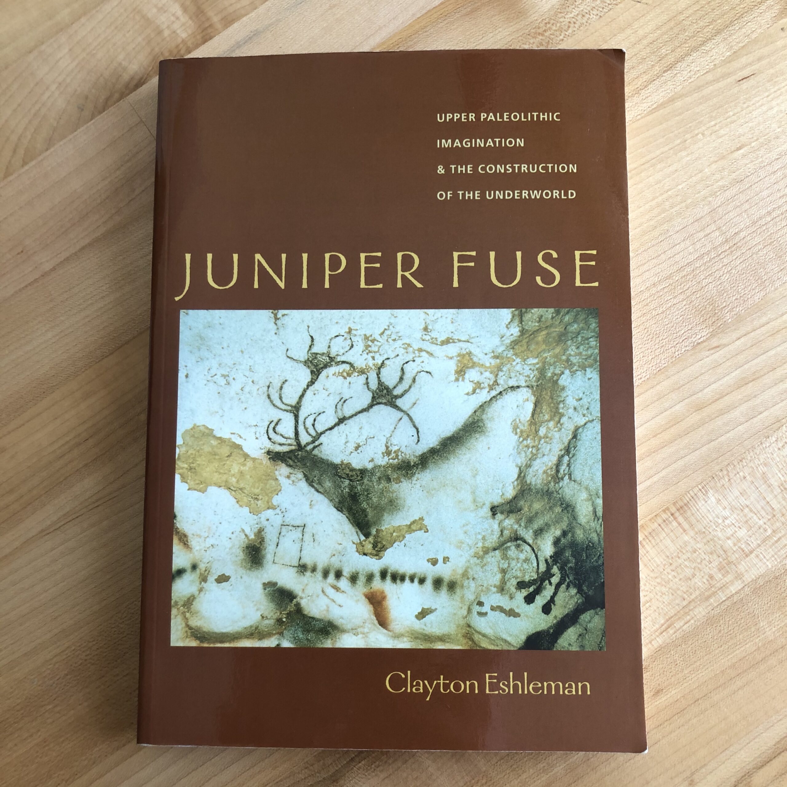 Juniper Fuse by Clayton Eshleman in paperback, lying on a maple tabletop. The brown bookcover features tan typography and a photo of a paleolithic cave painting.