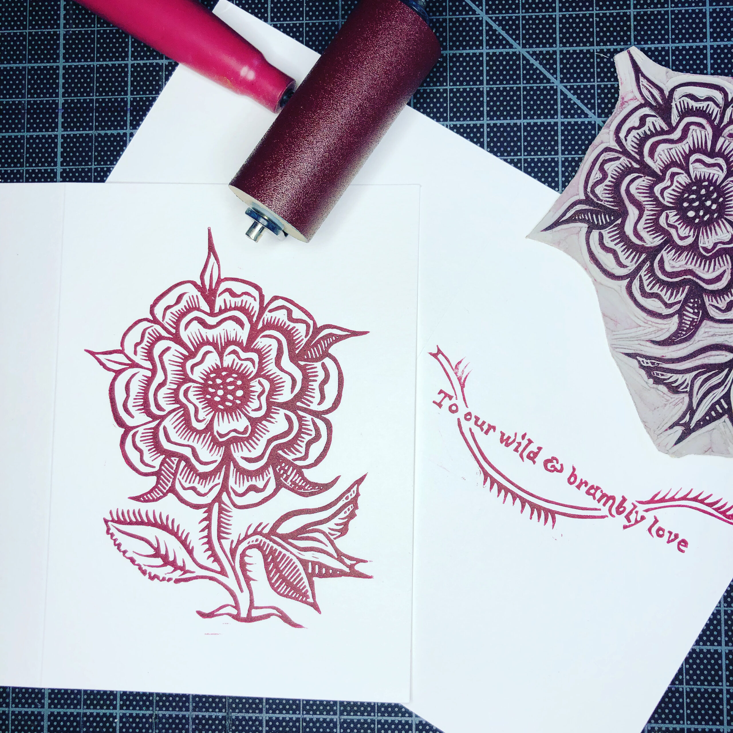 Handprinted linocut greeting card in red-violent ink on white paper, featuring a wild rose from a 16th-century book. A second card to the right shows hand-carved lettering and thorny stem inside reading: To our wild & brambly love. The linocut block and an inked roller are lying alongside the cards on a black gridded cutting mat.
