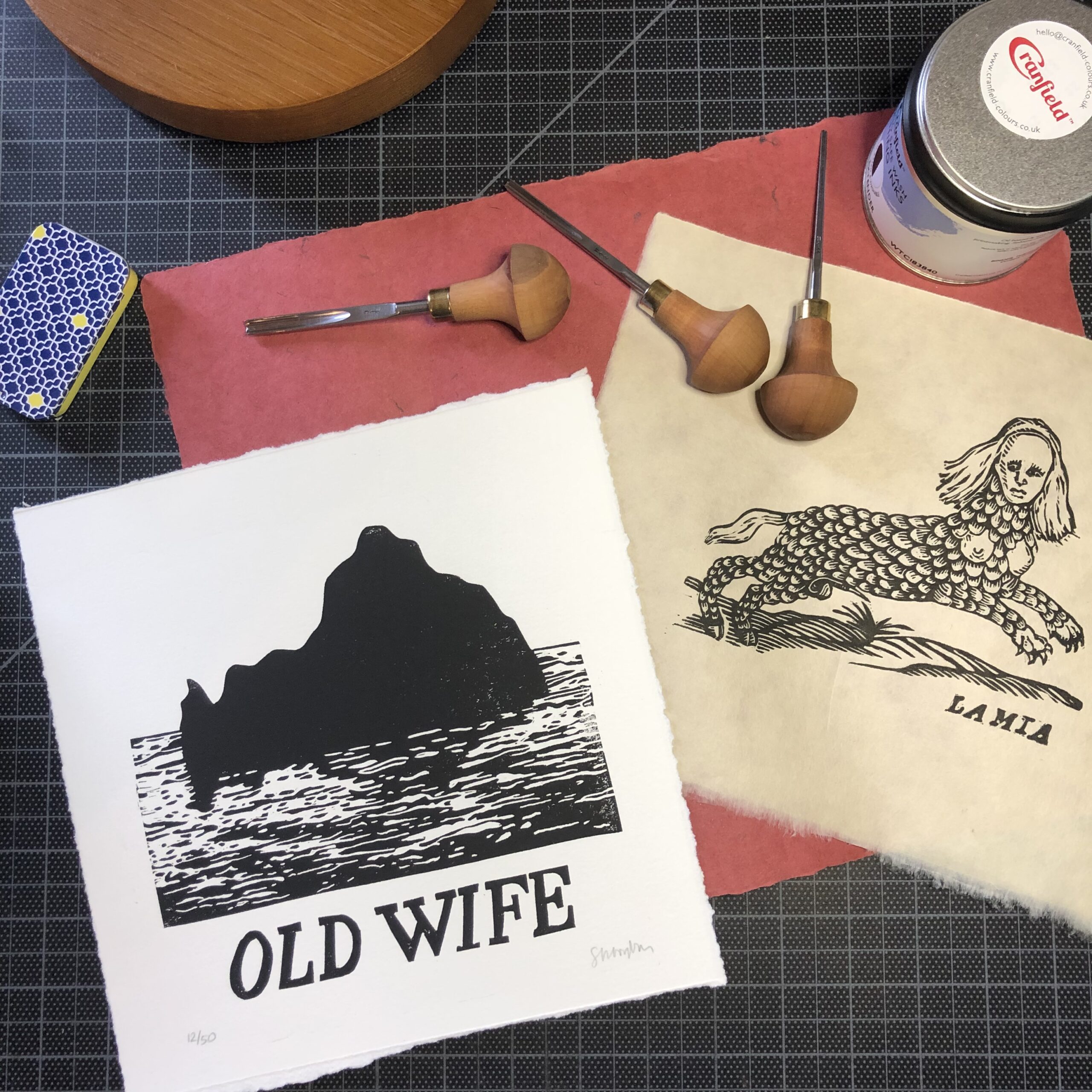 The Lamia linocut print lies on a workbench with carving tools and a jar of Cranfield Colors Safe Wash Ink. The Old Wife linocut print is to the left.