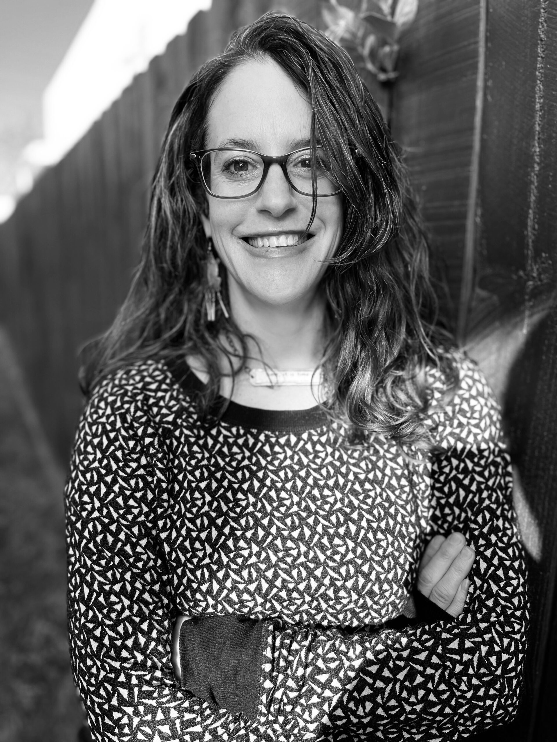 Reagan Louise Wilson in a black and white photo taken outside in front of a fence. She is smiling, wearing glasses and a patterned top.
