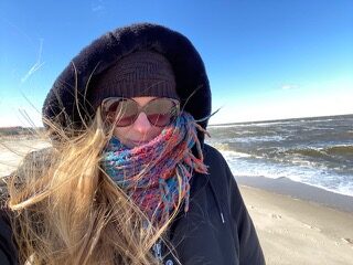Pattie McCarthy in a coat, hat, scarf, and large sunglasses stands on a beach in winter with her long blond hair whipping around her in the wind.