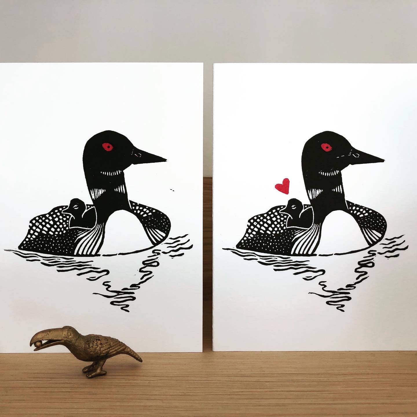 Two loon and chick cards side by side on a shelf. The left card has no heart above the chick, the right card does have a heart. Both cards show the adult loon with a red eye.