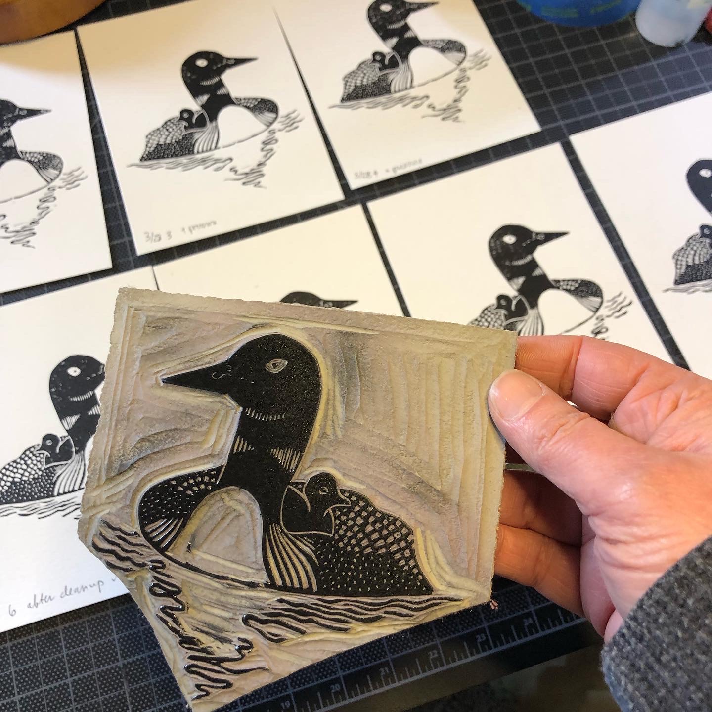 Several loon cards in progress arrayed on a table. In the foreground Shanna’s hand is holding the linked linoleum block from which the cards are printed.