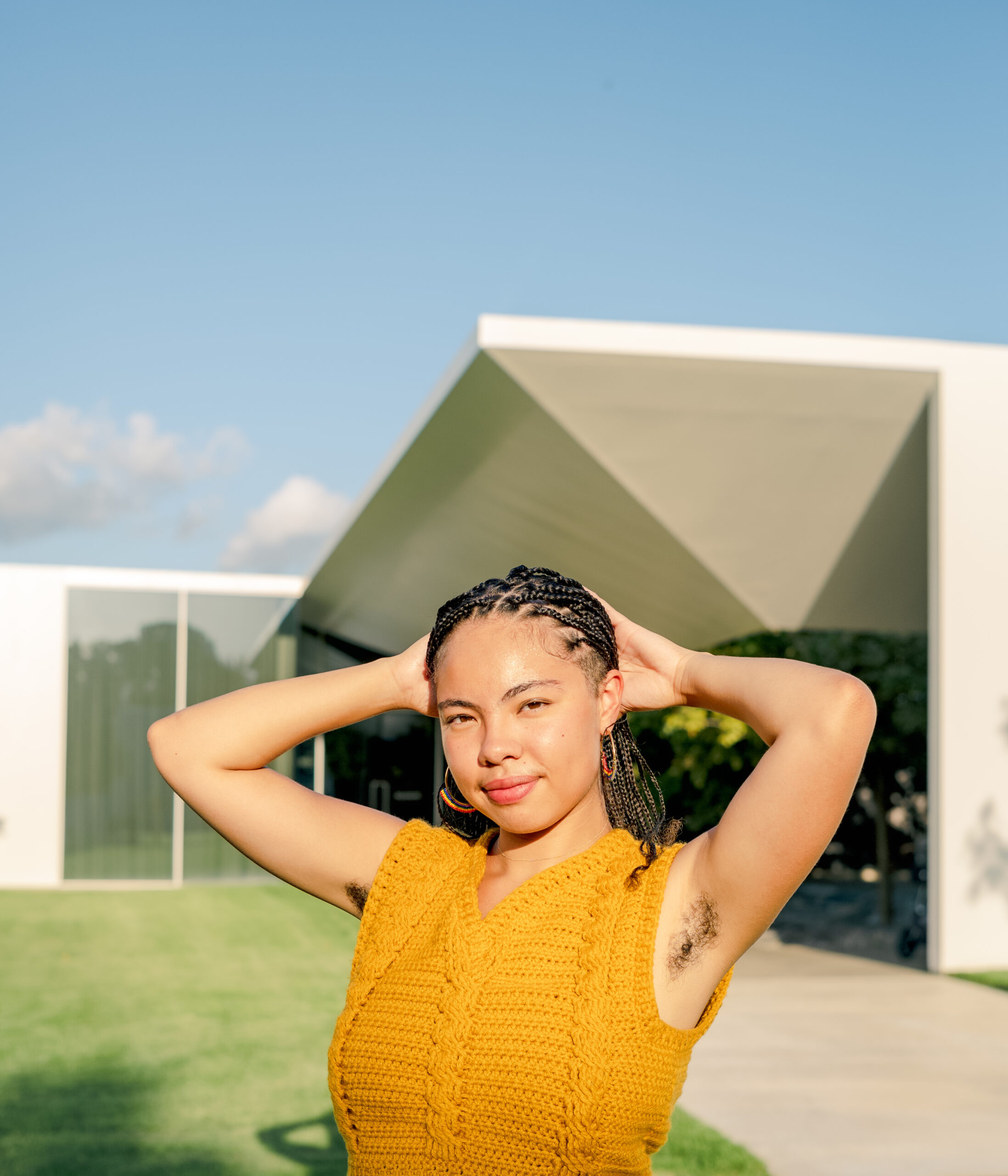 Irene Vázquez in a sleeveless yellow sweater in front of a modern building. Her hair is in braids and her arms are lifted with her hands behind her head.