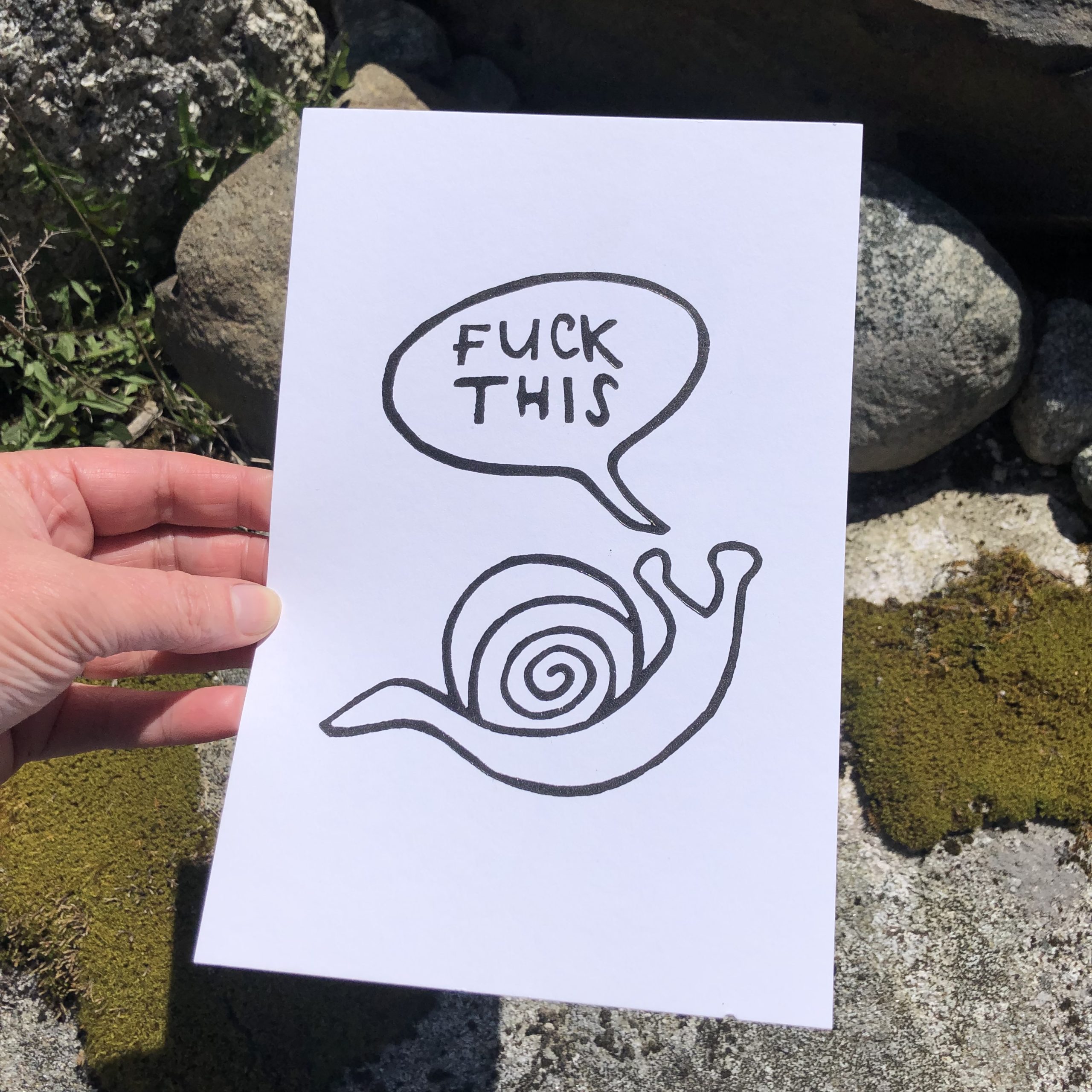 A white 6 x 9 linocut print held by a woman’s hand in front of some mossy boulders. The image printed in black ink is of the outline of snail with a speech bubble saying Fuck This in all caps.