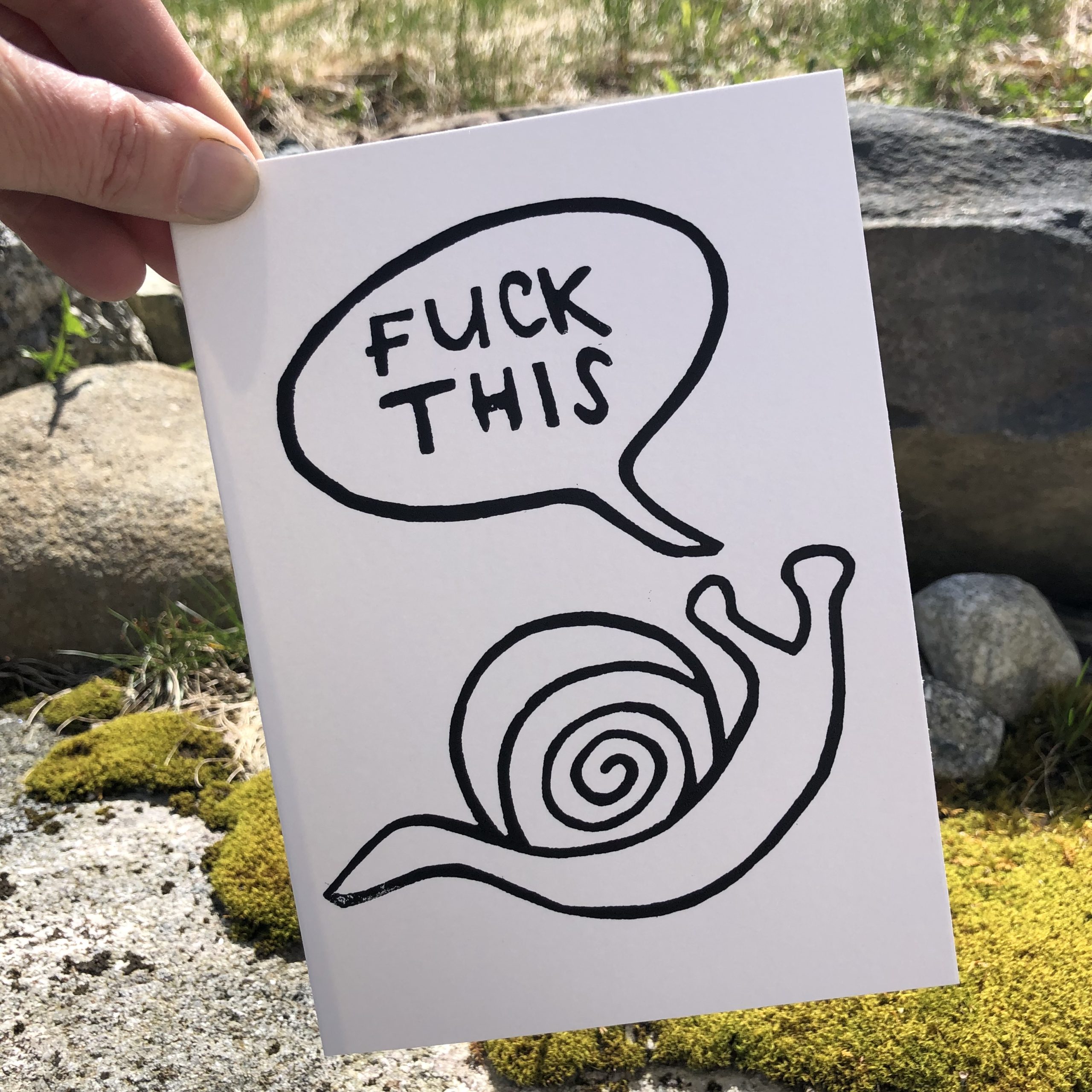 A white card with a black outline of a snail in blank ink. A speech bubble above the snail says “fuck this” in all caps. The style is low-fi and minimalist. The card is held by the upper left corner in a woman’s hand, outdoors with moss-covered boulders behind it.